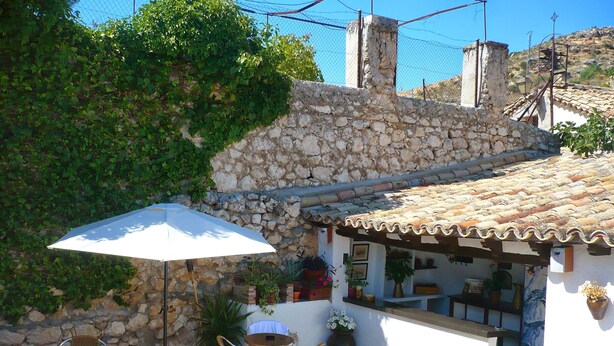 An outdoor patio with stone walls, a tiled floor, and a white umbrella. There are wicker chairs and a small table.