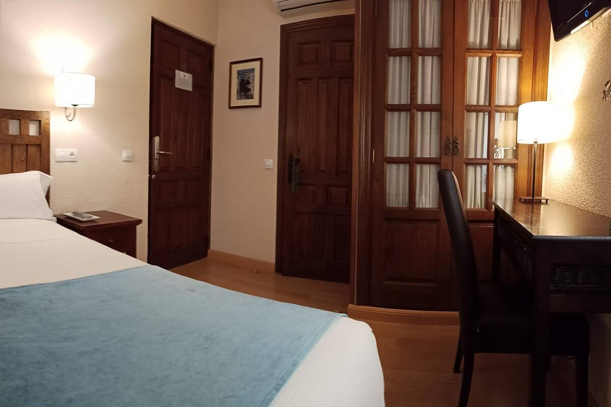 Quarto de hotel com cama, mesa de cabeceira, abajur e escrivaninha com cadeira.