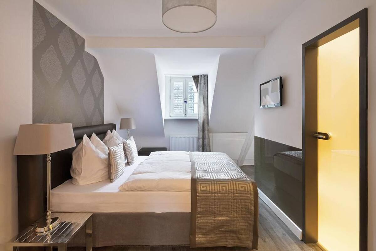Quarto de hotel moderno com uma cama grande, mesa de cabeceira, luminária, vista para a cidade e uma porta deslizante.