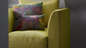 Un canapé jaune avec des coussins à motifs, dans une pièce dotée d’un plafond en pente et d’une fenêtre.
