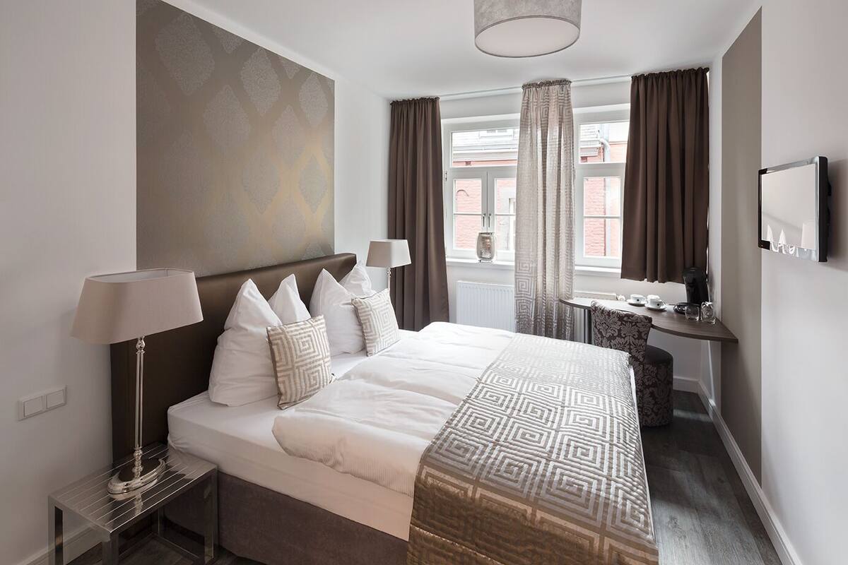 Quarto de hotel moderno com uma cama grande, luminárias de cabeceira, uma mesa de trabalho com cadeira e televisão na parede.