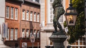 Une statue représentant un homme vêtu d’un costume d’époque, placée sur un piédestal, avec un lampadaire et des bâtiments en arrière-plan.