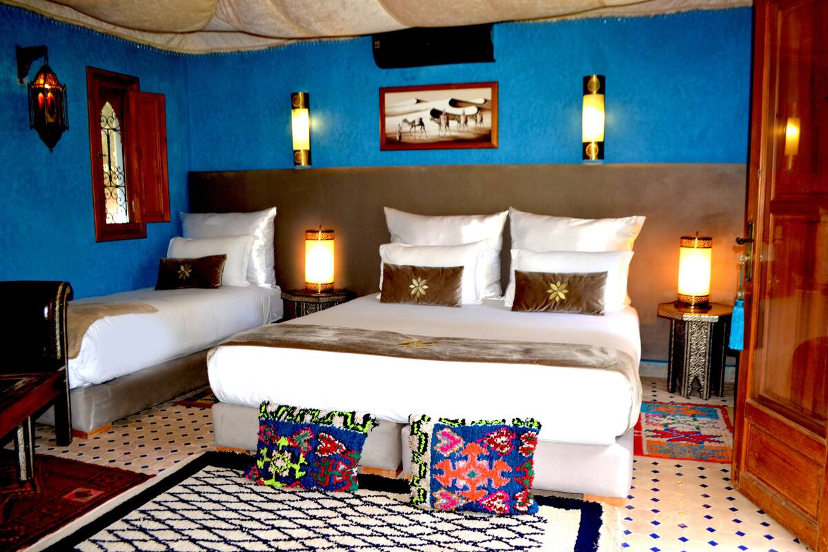 A hotel room with a large bed, two bedside tables, a nightstand, a wooden wardrobe, and a blue wall with a framed picture.