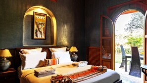A hotel room with a large bed, two bedside lamps, a painting on the wall, and a view of the outdoors through an open door.