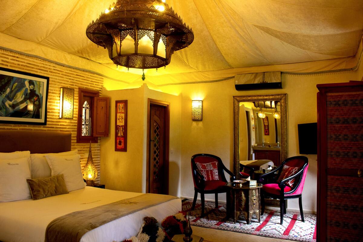 A bedroom with a large bed, two chairs, a small table, a chandelier, and a mirror.