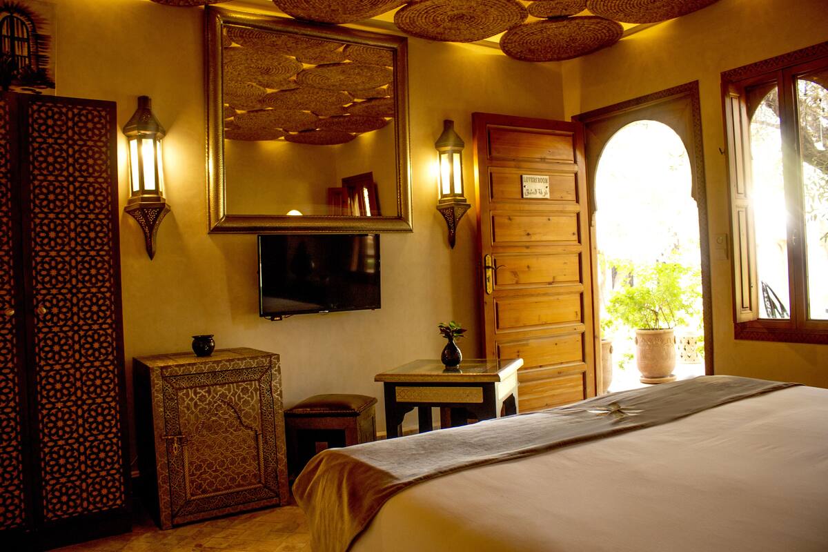 A hotel room with a bed, a television, wall sconces, a wooden wardrobe, a small table with a vase, and a large window with a view of greenery.