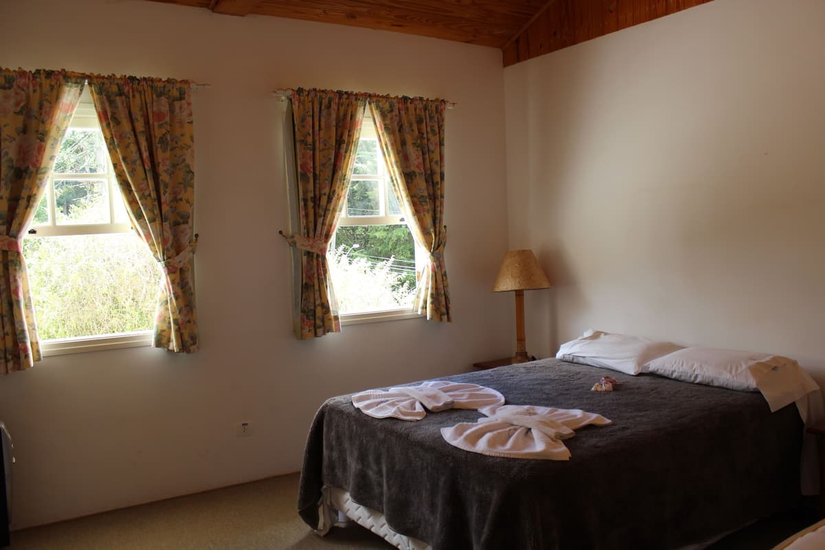 Ein Schlafzimmer mit zwei Fenstern, einem Bett mit dunkler Bettdecke und einer Nachttischlampe.