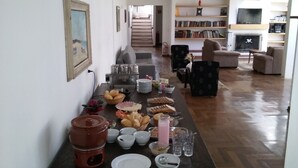 A dining table set with various food items and drinks, a bookshelf in the background, and a living room area with a sofa and chairs. - Pousada Vila Menotti (Campos do Jordão)