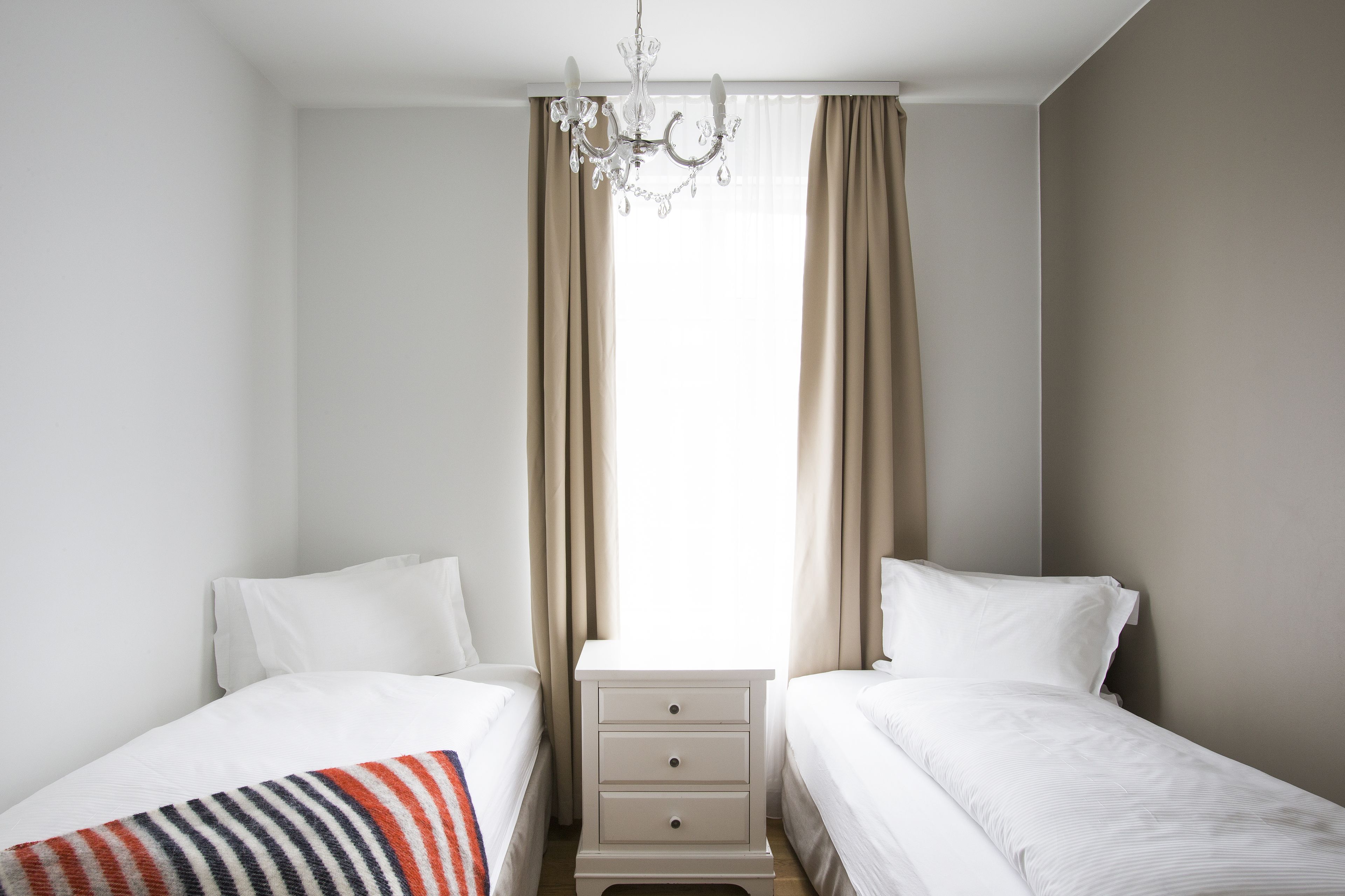 A bedroom with two beds, a nightstand, a chandelier, and beige curtains.