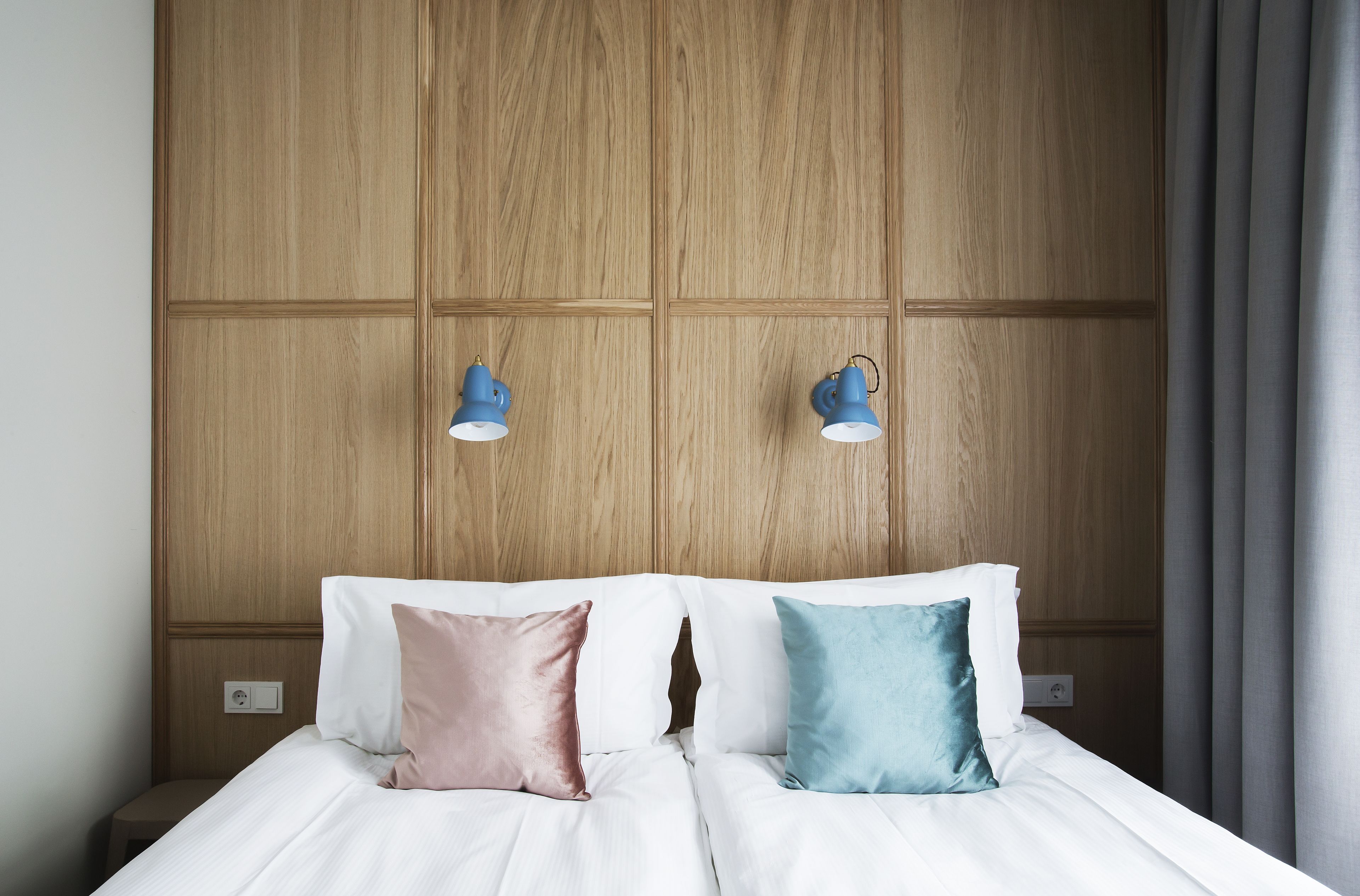 Quarto de hotel com cabeceira de madeira, duas luminárias de cabeceira e cama com roupa de cama branca e dois travesseiros.