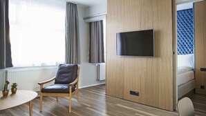 Une chambre d’hôtel moderne avec un séparateur en bois, un téléviseur à écran plat, un lit avec une tête de lit bleue, une chaise en bois, une petite table avec des plantes et une fenêtre avec des rideaux.