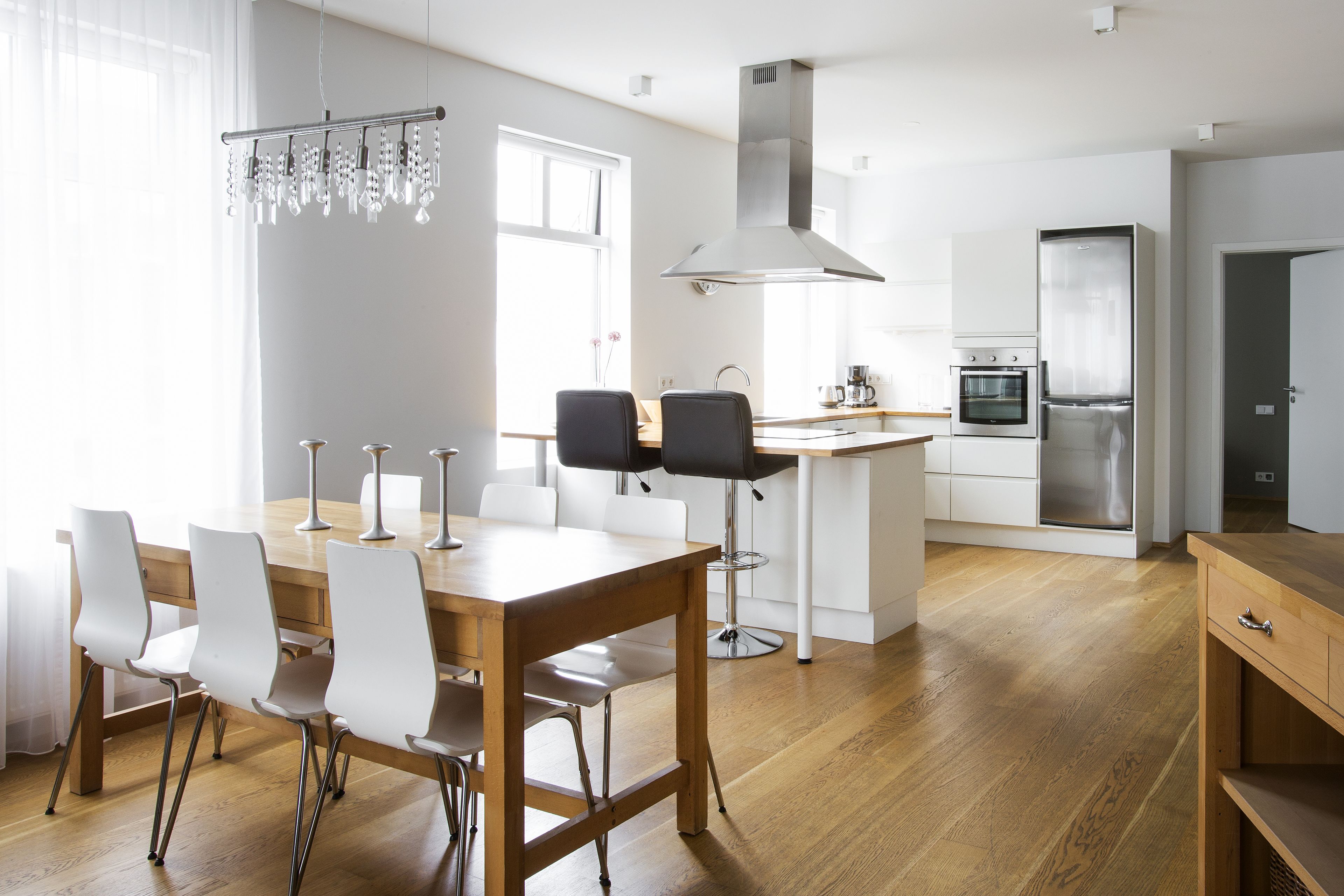 A modern kitchen with a wooden dining table, white chairs, stainless steel appliances, and a hanging light fixture.