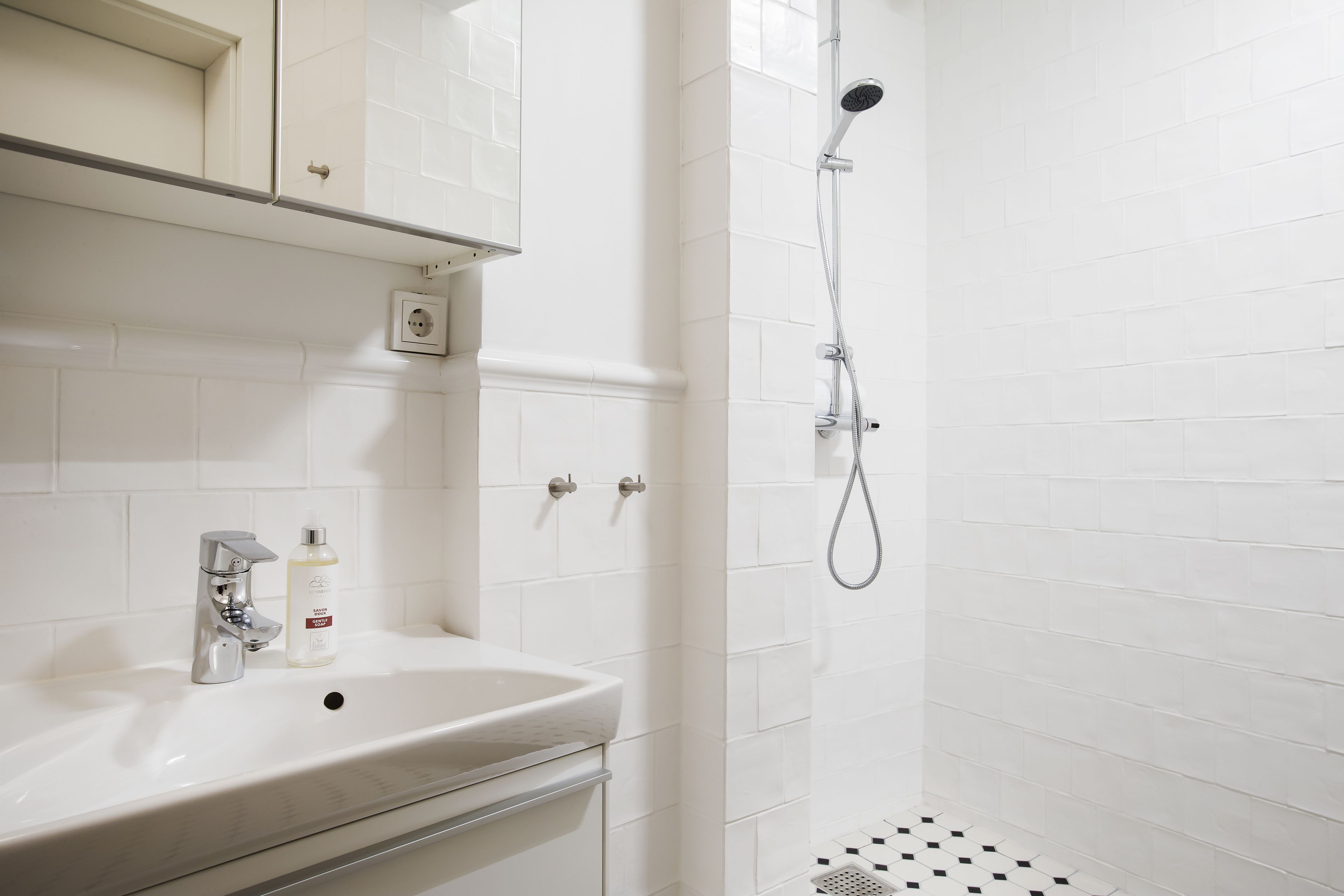 Un baño con lavamanos blanco, espejo, ducha y piso de baldosas.