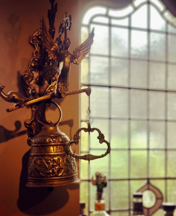 A brass bell with intricate designs and a figure, hanging in front of a window with a grid pattern. - The Wheatsheaf (Milnthorpe)