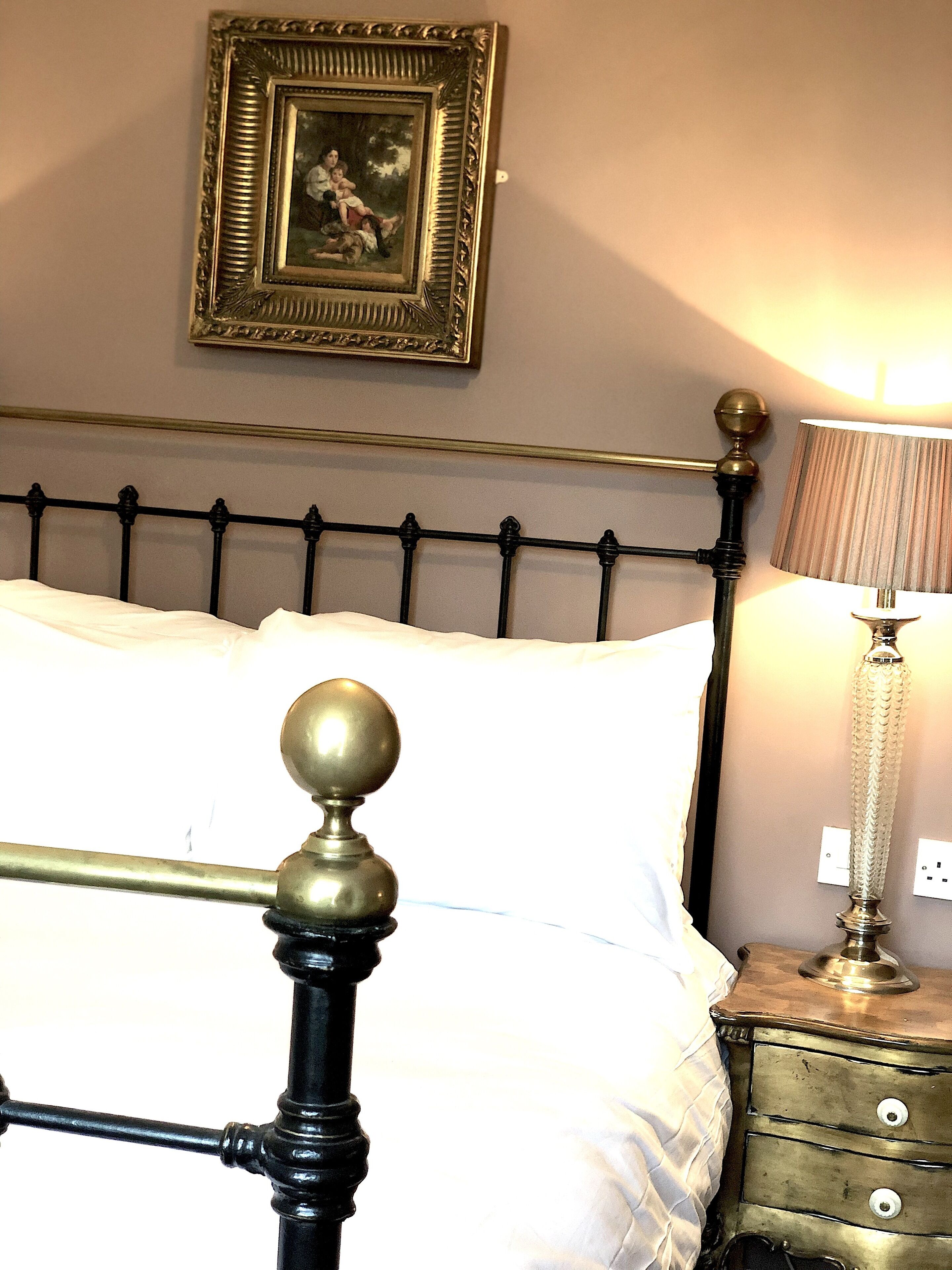 Una cama con estructura de metal negro y detalles en latón, una mesita de noche con una lámpara, y un cuadro enmarcado en la pared.