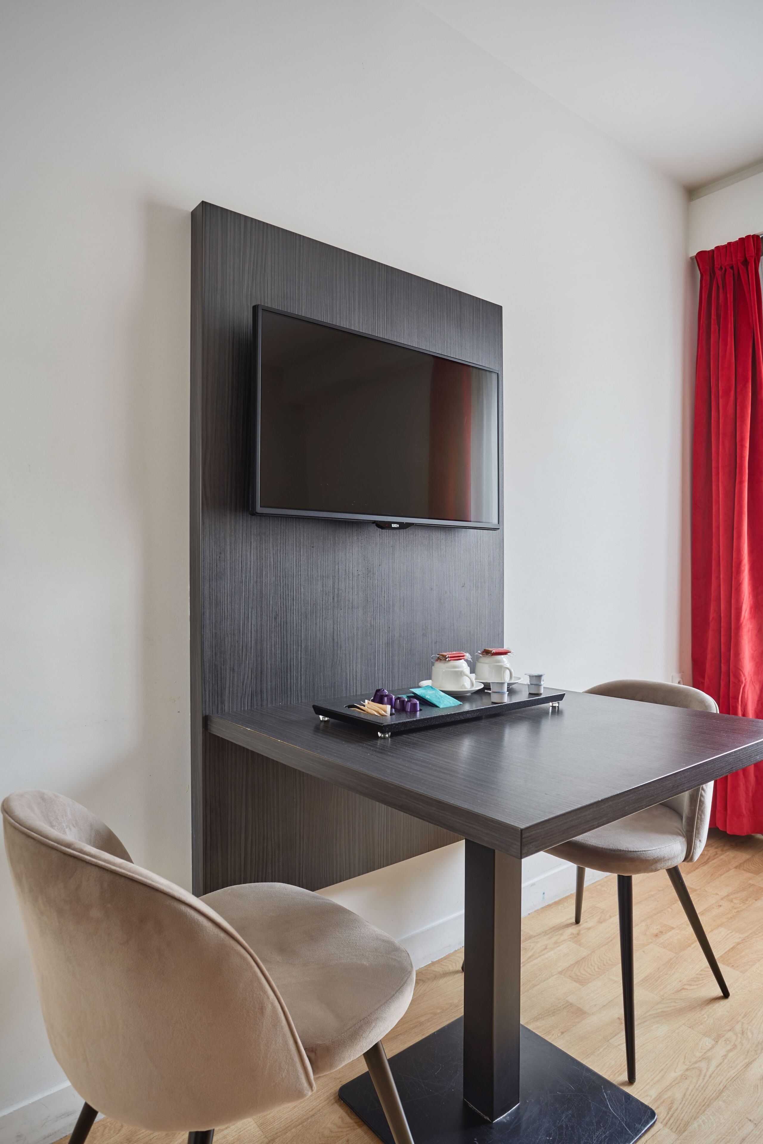 A modern room with a flat-screen TV mounted on a dark wall, a small table with a tray of items, two chairs, and a red curtain.