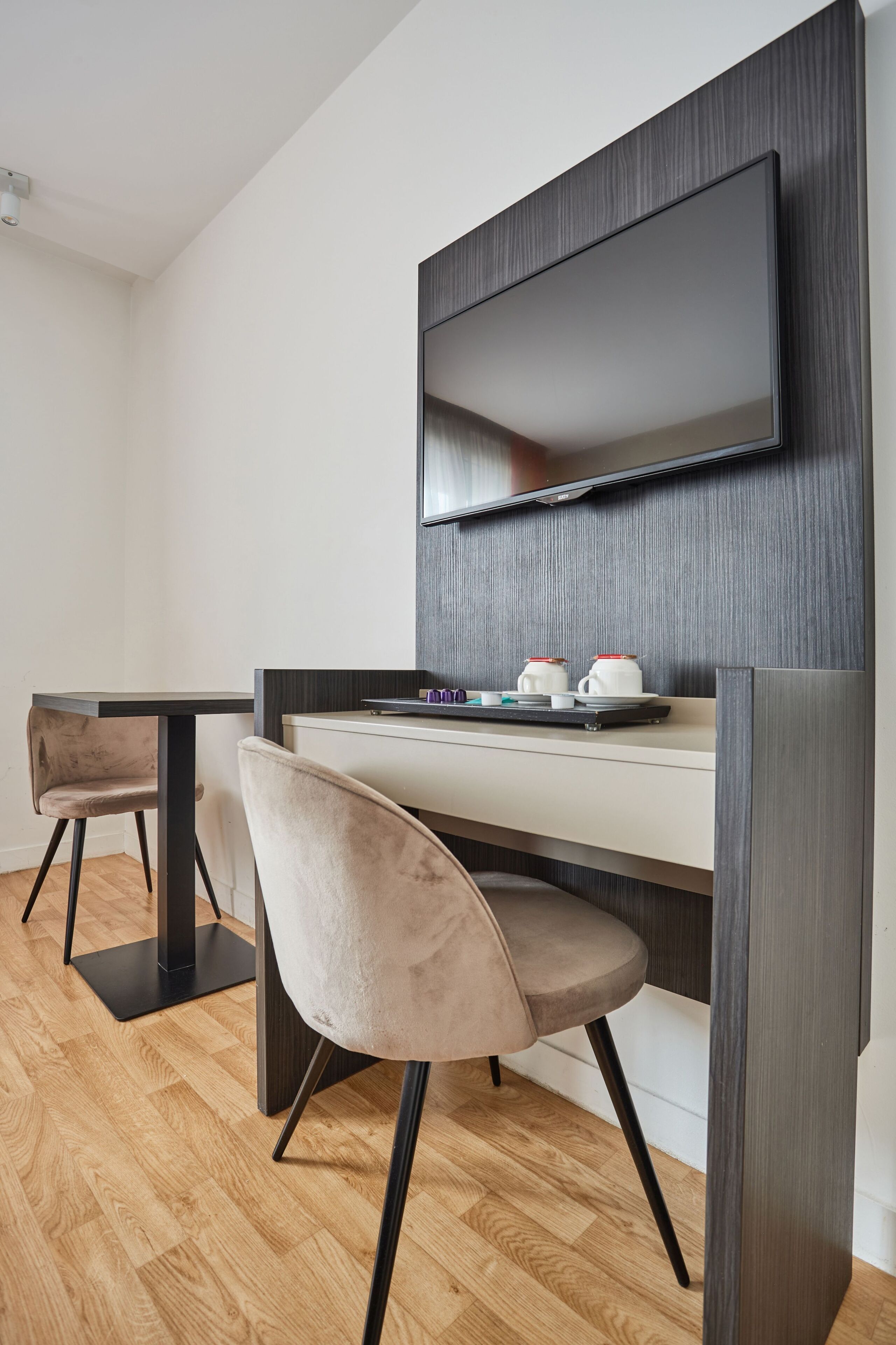 Una camera d'albergo moderna con pavimento in legno, una TV a schermo piatto montata a parete, un tavolino con due tazze e un vassoio, e due sedie.