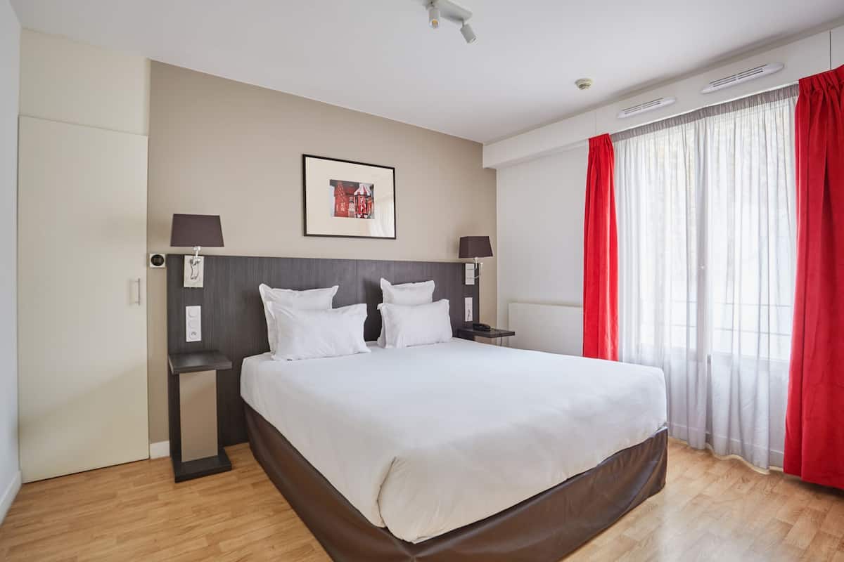 Une chambre d’hôtel avec un grand lit, deux tables de chevet, une tête de lit, un tableau encadré au mur et une fenêtre avec des rideaux rouges.