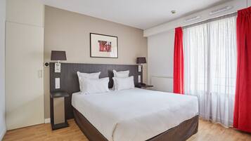 Una habitación de hotel con una cama grande, dos mesitas de noche, un cabecero, un cuadro enmarcado en la pared y una ventana con cortinas rojas.