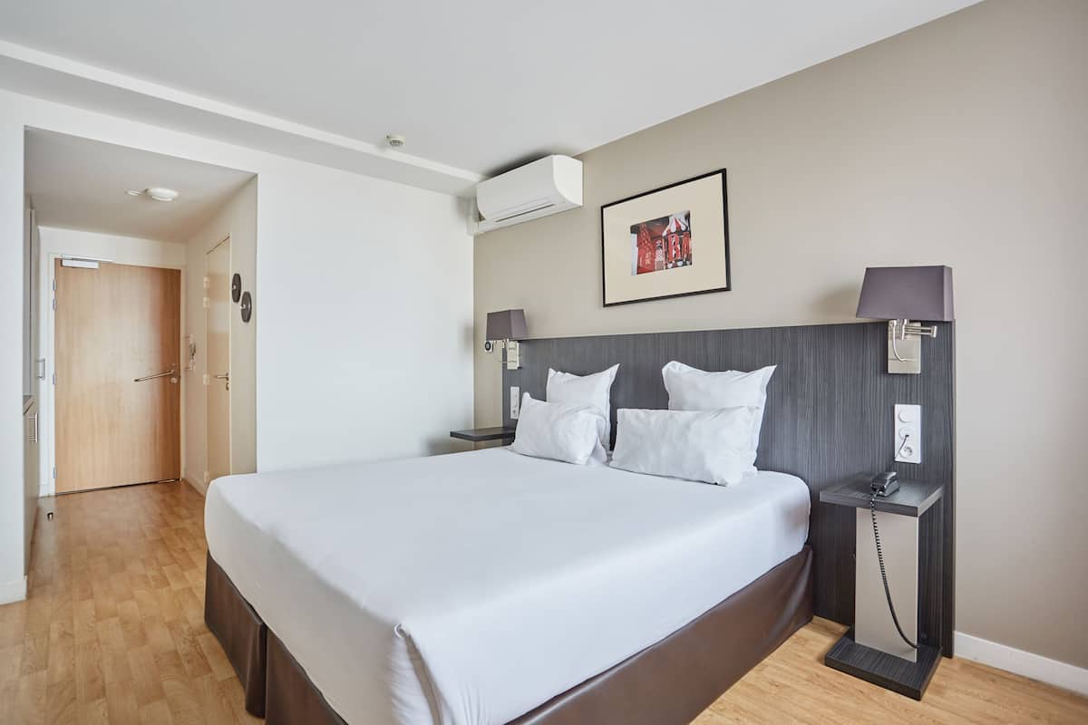Une chambre d’hôtel moderne avec un grand lit, une table de chevet avec une lampe, un climatiseur fixé au mur et un tableau encadré au-dessus du lit.