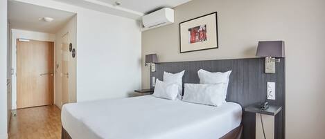 A modern hotel room with a large bed, a bedside table with a lamp, a wall-mounted air conditioner, and a framed picture above the bed.