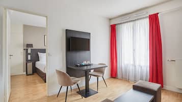 A modern hotel room with a flat-screen TV mounted on the wall, a small dining table with two chairs, a bench, and a large window with red curtains.