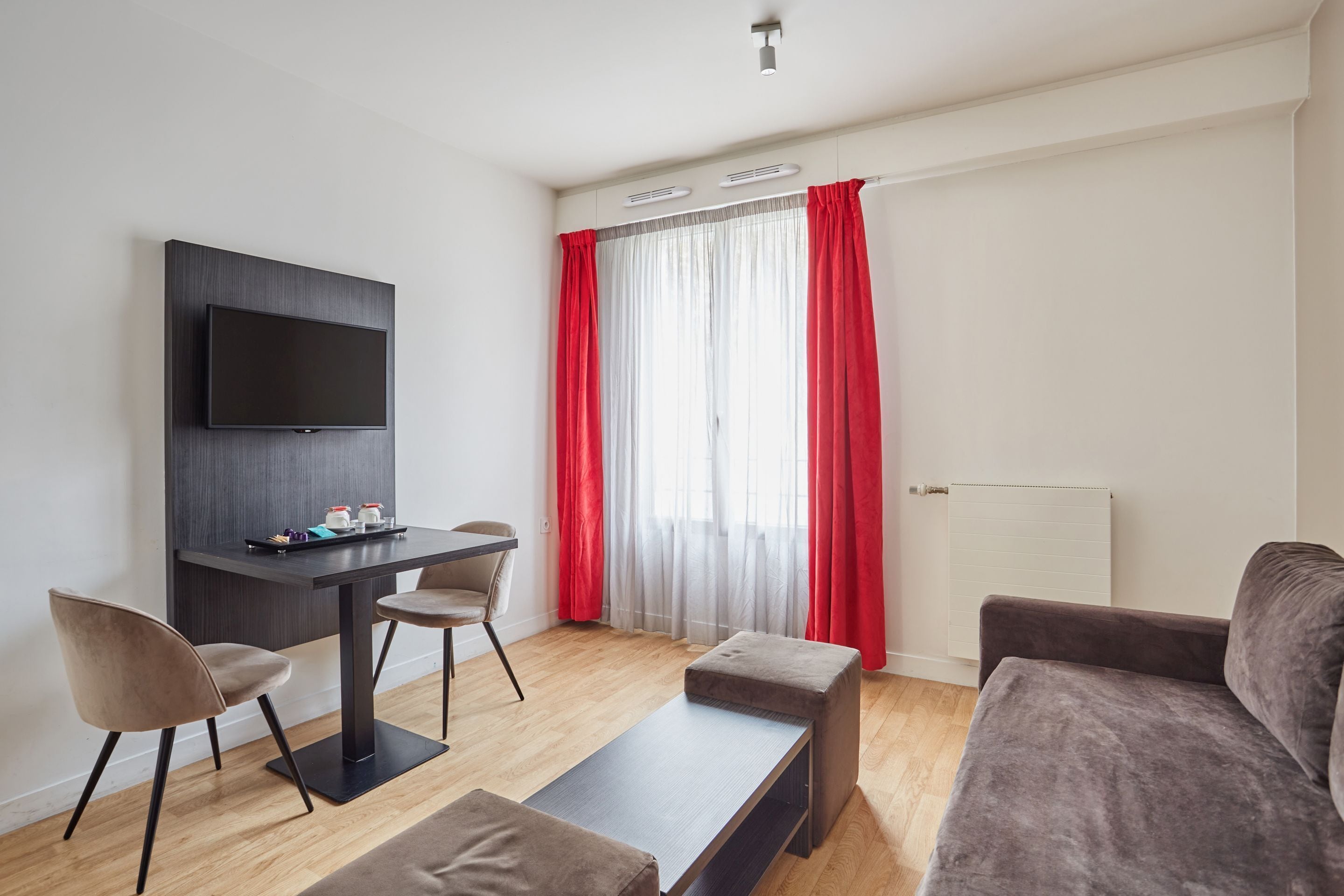 Un salon moderne avec un téléviseur à écran plat fixé au mur, une table en bois foncé avec deux chaises, un canapé gris et un rideau rouge qui recouvre une fenêtre.