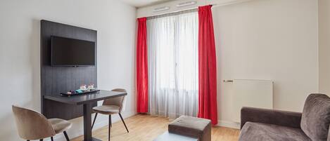 A modern living room with a flat-screen TV mounted on the wall, a dark wooden table with two chairs, a grey sofa, and a red curtain covering a window.