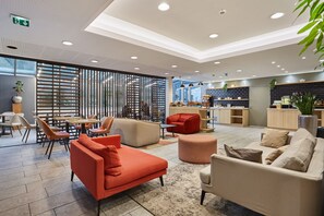 Een moderne hotellobby met zithoeken, een keuken op de achtergrond en een groot raam.
