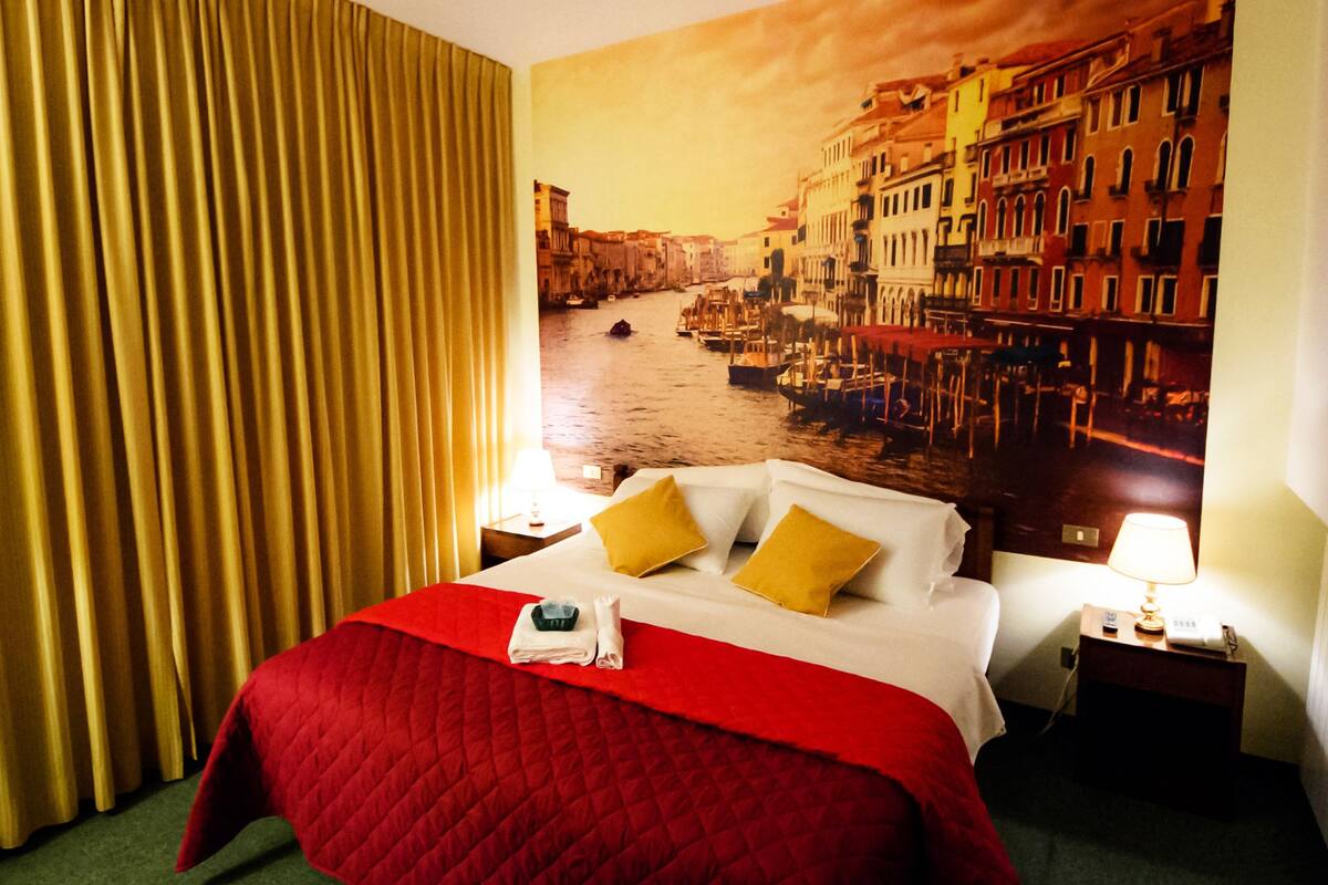 Ein Hotelzimmer mit Bett, Vorhängen, einem Nachttisch mit Lampe und einem Wandbild einer Stadtansicht.