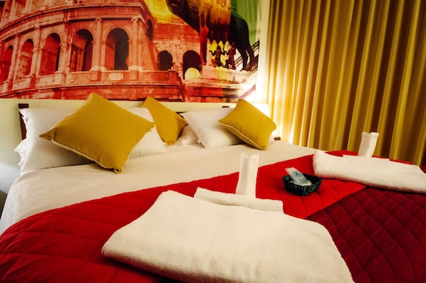 A hotel room with a bed featuring red and white bedding, multiple pillows, and neatly folded towels.