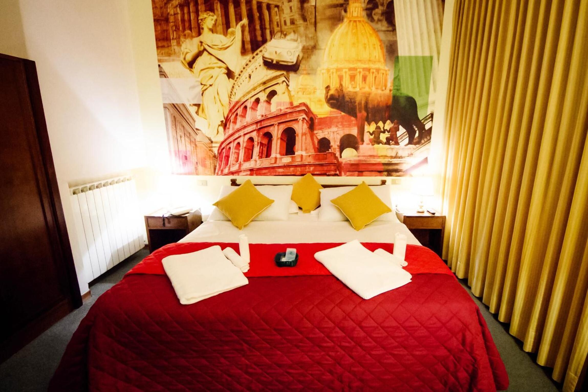 A hotel room with a large bed, red and yellow pillows, a red quilted bedspread, and a wall mural depicting a cityscape.