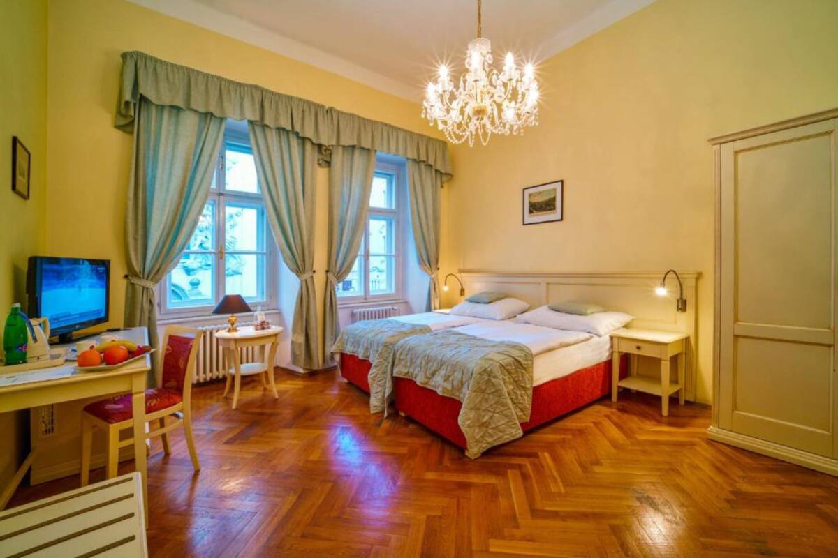 A bedroom with a wooden floor, a bed with red bedding, a desk with a computer, a chair, and a chandelier.
