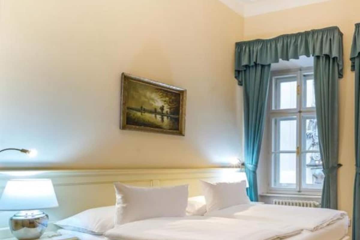 A neatly made bed in a room with a wooden floor, a nightstand with a lamp, a painting on the wall, and a window with curtains.