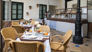 Un comedor con sillas y mesas de mimbre dispuestas para el desayuno, una cocina al fondo y un pilar decorativo.