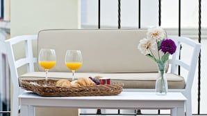 Un área de estar al aire libre con mobiliario blanco y una cesta de mimbre que contiene croissants, un botellón de jugo y un pequeño recipiente, probablemente yogur o crema.