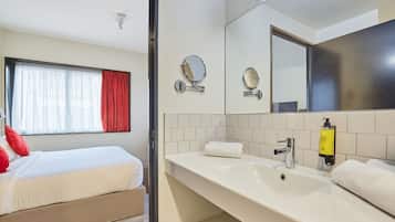 Ein Hotelzimmer mit Bett, Bad mit Waschbecken, Spiegel und Fenster mit roten Vorhängen.