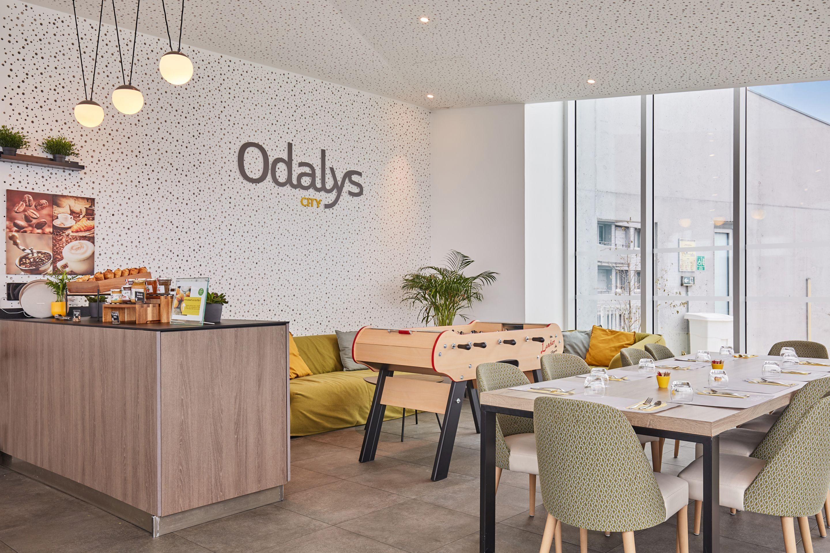 Un intérieur moderne avec un comptoir d’accueil en bois, une table de baby-foot et un coin repas doté de chaises à motifs verts.