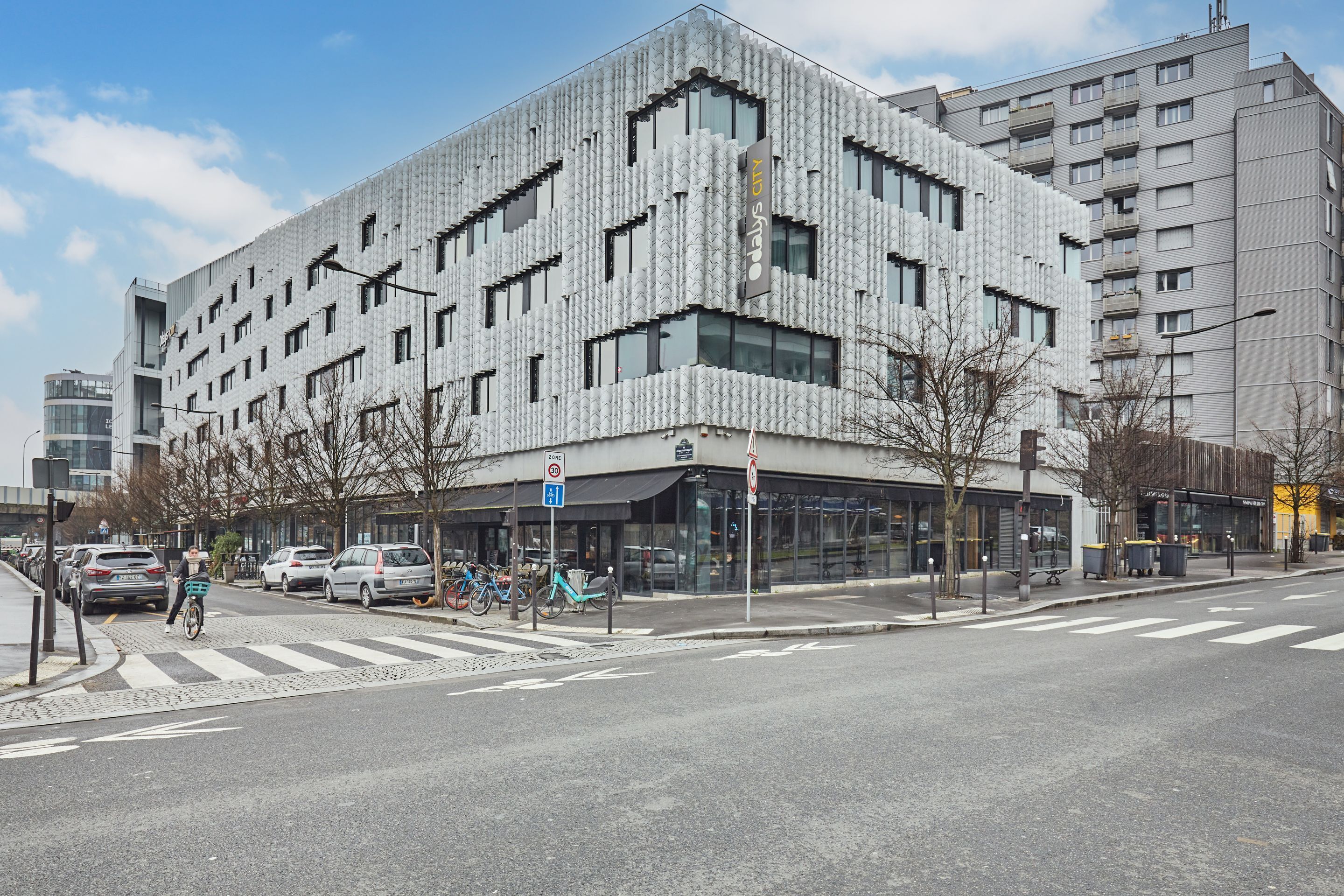 Un bâtiment moderne doté d’une façade originale, d’un passage piéton et de voitures garées.