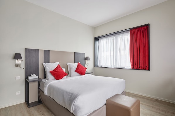 A modern hotel room with a large bed, two bedside tables, a nightstand, a window with red curtains, and a wooden floor. - Appart Hotel Odalys City Paris XVII (Paris)