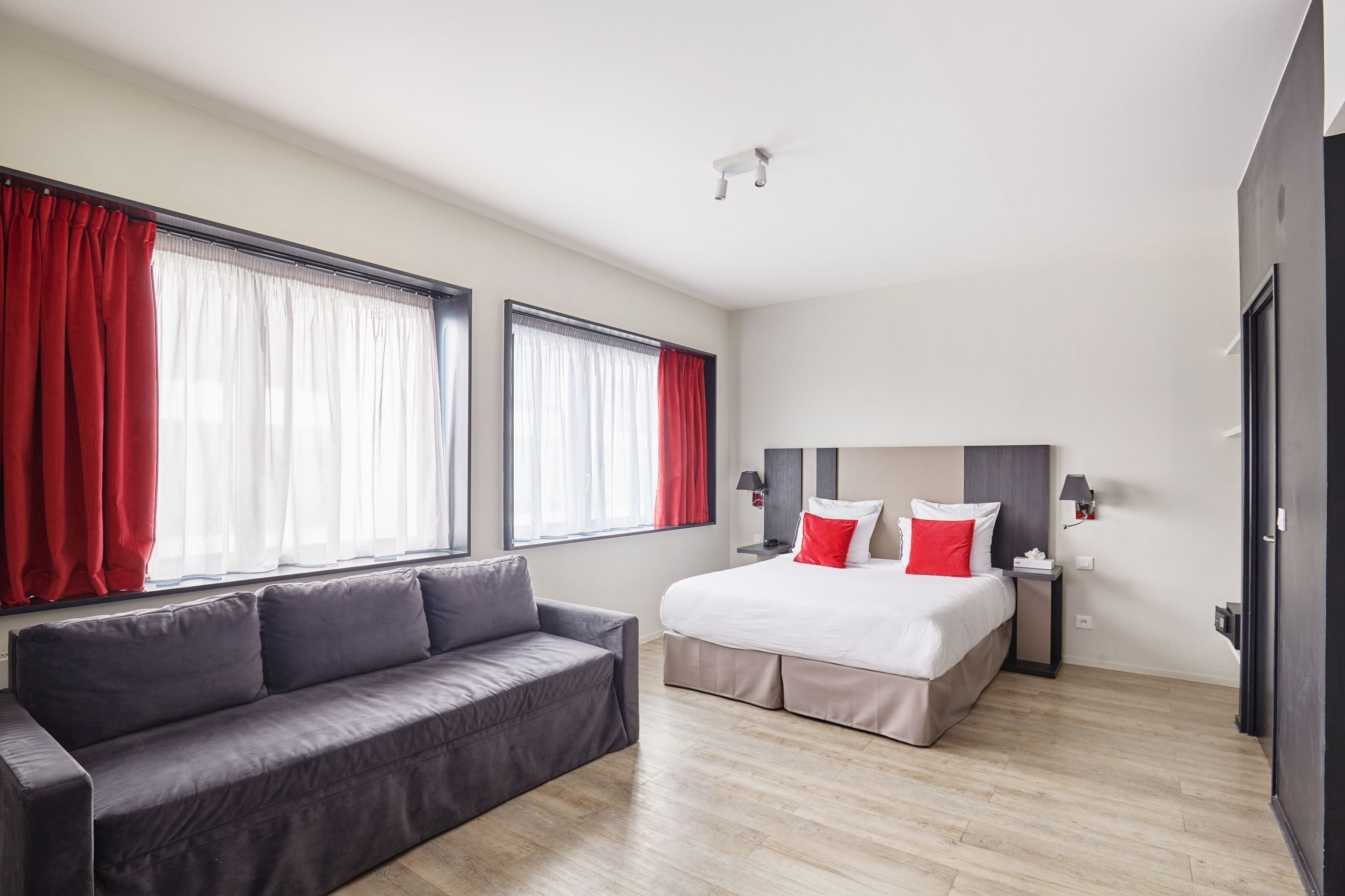Une chambre d’hôtel moderne avec un grand lit, un canapé gris et deux fenêtres avec des rideaux rouges.