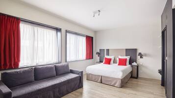 Une chambre d’hôtel moderne avec un grand lit, un canapé gris et deux fenêtres avec des rideaux rouges.