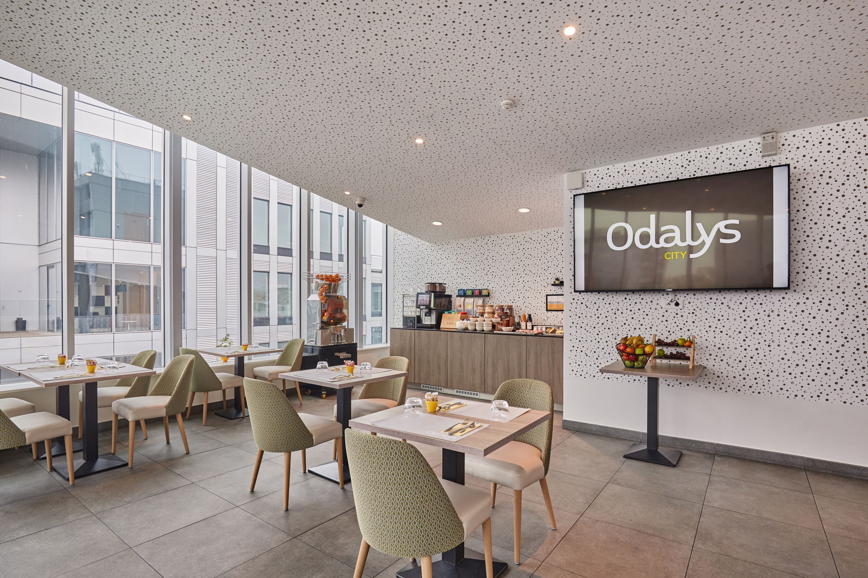 Un comedor moderno con mesas y sillas, una pantalla grande mostrando 'Odaly's City', y una zona de cocina al fondo.