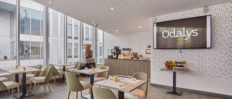 Un comedor moderno con mesas y sillas, una pantalla grande mostrando 'Odaly's City', y una zona de cocina al fondo.
