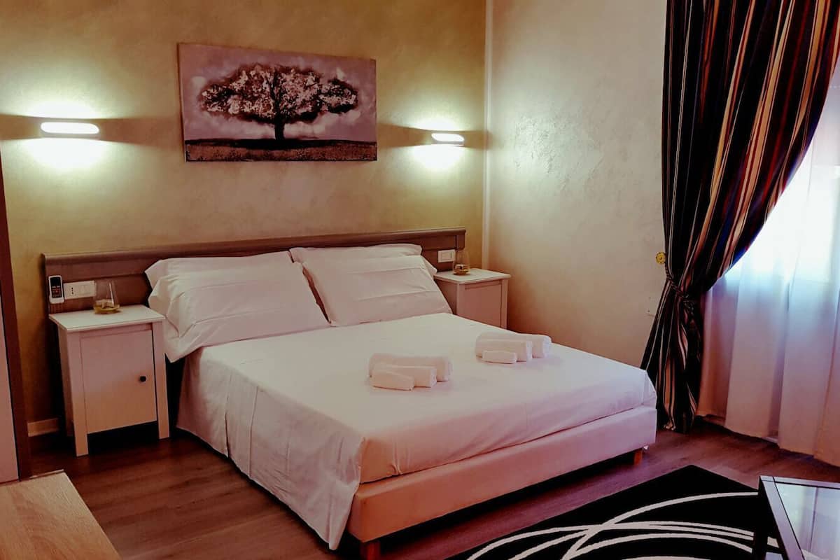 A neatly made bed with white linens and two rolled towels, a bedside table with a lamp, a wall-mounted artwork of a tree, and a window with curtains.