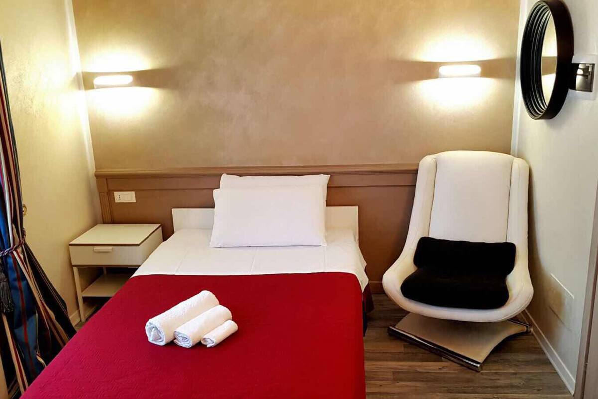 A hotel room with a bed, a chair, a nightstand, and a wall-mounted light fixture.