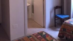 Un dormitorio con cama, una silla, televisor montado en la pared y una puerta que da a un baño.