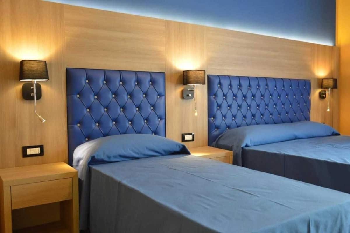 Een hotelkamer met twee bedden, elk met een blauw hoofdeinde en bijpassende kussens. Er staan nachtkastjes met lampen en er is een wandlamp.