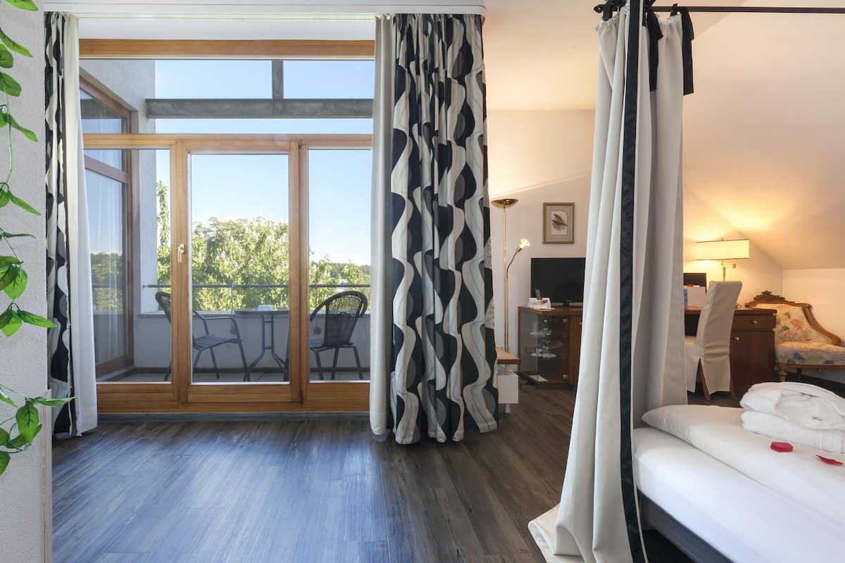 Habitación de hotel con balcón, cama con ropa blanca, escritorio con silla y lámpara.