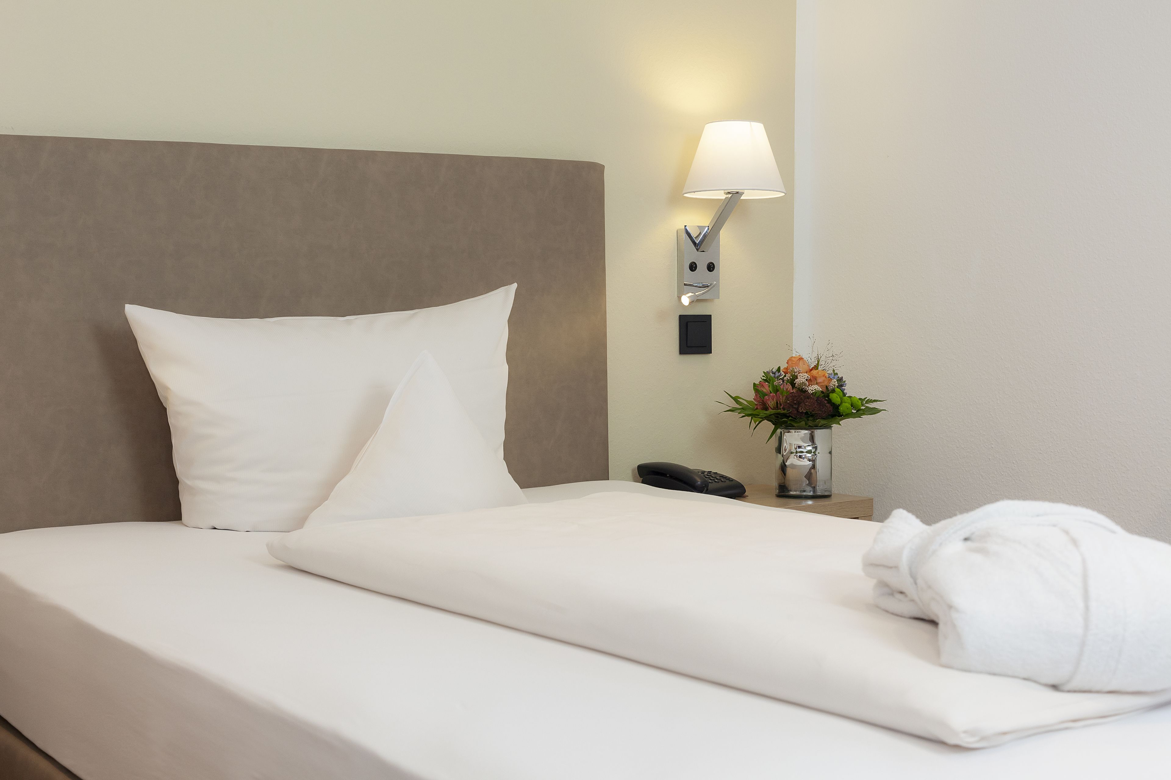 Ein ordentlich bezogenes Bett mit weißen Leinentüchern, eine Nachttischlampe, ein Telefon und eine Vase mit Blumen.